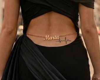 May include: A black floor-length gown featuring an off-the-shoulder neckline with draped fabric and a large back cutout. A gold-toned chain waist accessory is visible, featuring the name Maria in script font and a heartbeat line graphic.