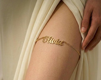 May include: A gold-toned metal thigh garter featuring a delicate chain band and a central nameplate with the script text Olivia. The piece is designed to encircle the upper leg, showcasing a polished finish and a cursive font for the name.