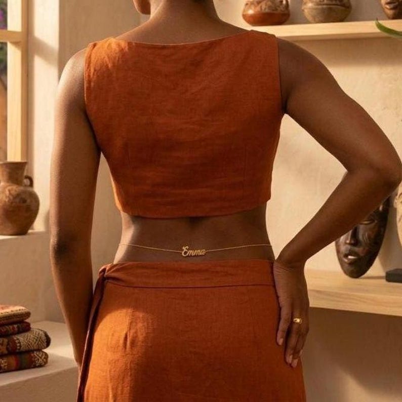 May include: A rust-colored sleeveless crop top and matching skirt set featuring a thin gold-toned waist chain with the name Emma in cursive script. The garment has a textured fabric finish and is worn with a simple gold band on the finger.