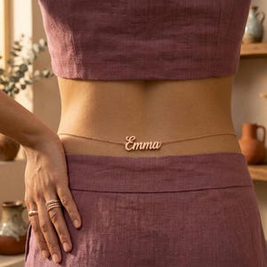 May include: A rose gold-toned waist chain featuring a delicate cable link chain and a central nameplate pendant with the name Emma written in a cursive script font. The jewelry piece is worn around the midsection over a mauve-colored linen garment.
