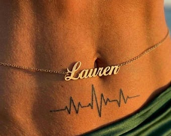 May include: A gold-toned metal belly chain featuring a central nameplate with the name Lauren written in a cursive script font. The delicate chain links extend horizontally across the midsection, resting above a black ink heartbeat tattoo.