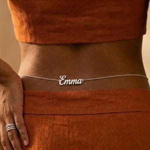 May include: A silver-toned belly chain featuring a central nameplate with the name Emma written in a cursive script font. The delicate metal chain connects to the nameplate on both sides, creating a continuous piece of jewelry worn around the waist.