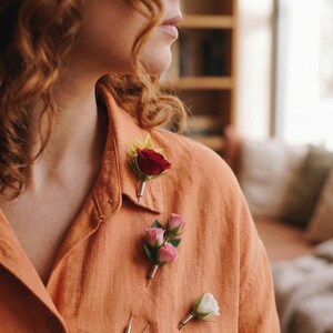 May include: An orange linen shirt adorned with three floral boutonnieres. One boutonniere features a single red rose, another has three pink roses, and the third has a single white rose. Several silver pins are also visible.
