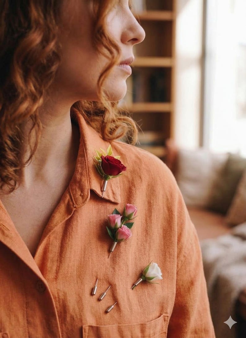 May include: An orange linen shirt adorned with three floral boutonnieres. One boutonniere features a single red rose, another has three pink roses, and the third has a single white rose. Several silver pins are also visible.