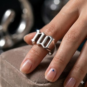 May include: A silver ring with the word "NO" in bold, block letters. The ring is worn on a finger, and the design is modern and eye-catching. The ring is made of a shiny, reflective metal.