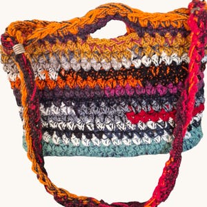 Op de afbeelding: Handgemaakte gehaakte tas met diverse kleuren, waaronder oranje, paars, grijs, wit en blauwgroen. De tas heeft een gevlochten schouderriem en een handvat aan de bovenkant. Het ontwerp heeft een gestructureerd steekpatroon en een klein rood accent.