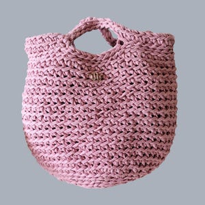 May include: A dusty rose-colored, hand-crocheted tote bag. The bag features a woven design with two handles and a small metal emblem. The bag's texture is visible, and the overall shape is round. The bag is suitable for carrying various items.