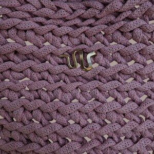 May include: Close-up of a woven, dusty rose fabric with a repeating braid pattern. A silver-colored metal snake emblem is affixed to the fabric. The texture of the fabric is visible, with a slightly open weave.