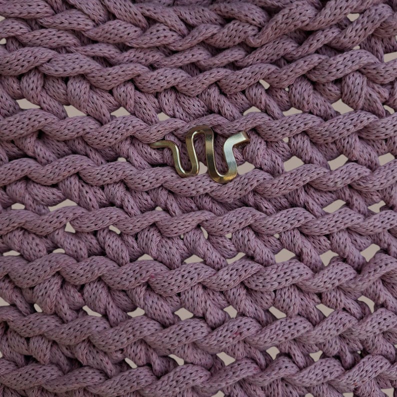May include: Close-up of a woven, dusty rose fabric with a repeating braid pattern. A silver-colored metal snake emblem is affixed to the fabric. The texture of the fabric is visible, with a slightly open weave.