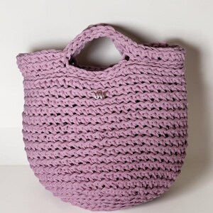 May include: A lavender-colored, handmade crochet tote bag. The bag features a rounded bottom, a sturdy handle, and a small silver-colored embellishment. The crochet stitch creates a textured surface. The bag is designed for carrying items.