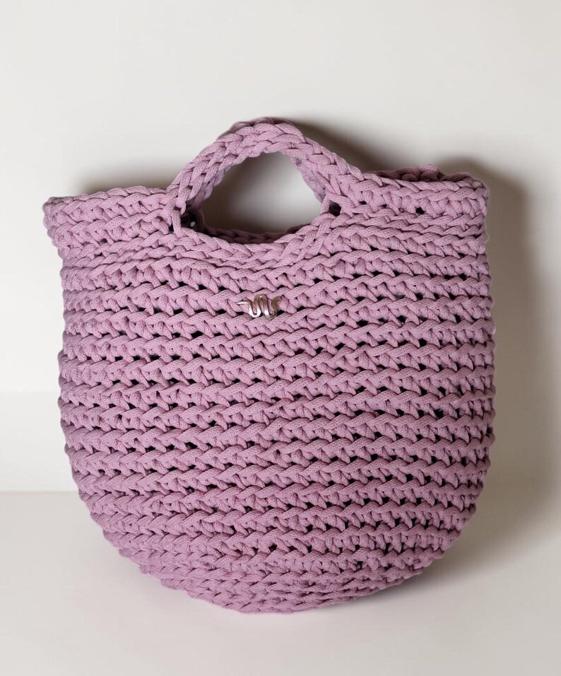 May include: A lavender-colored, handmade crochet tote bag. The bag features a rounded bottom, a sturdy handle, and a small silver-colored embellishment. The crochet stitch creates a textured surface. The bag is designed for carrying items.