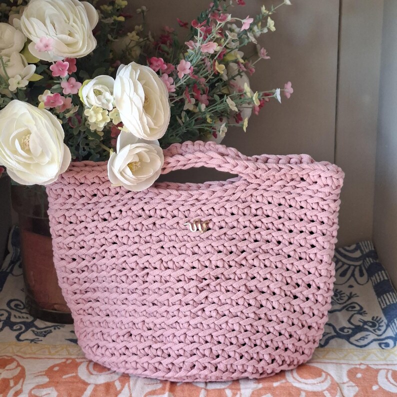 May include: A light pink, hand-crocheted tote bag with a woven texture. The bag has a small handle and a gold-colored metal emblem. White and pink flowers are in the background.