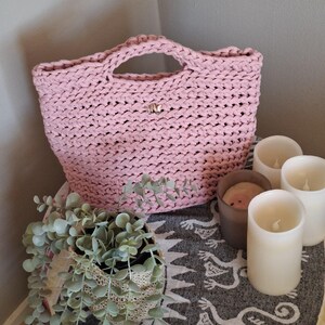 May include: A light pink, chunky-knit tote bag with two handles. The bag has a small, decorative silver clasp. The bag is displayed with a potted plant and several white pillar candles.