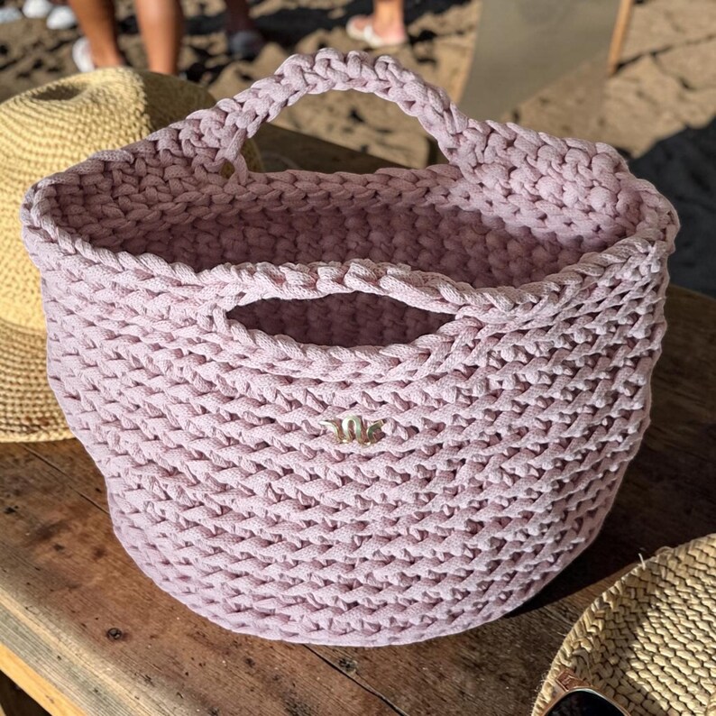 May include: A light purple, hand-knitted tote bag with a sturdy handle. The bag has a woven texture and a small gold-colored emblem on the front. The bag is sitting on a wooden surface. A straw hat is visible in the background.