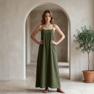 Puede incluir: Un vestido largo verde oliva con tirantes finos. El vestido tiene un escote cuadrado y un corte holgado y fluido. La modelo está de pie en un pasillo con arcos y un olivo en maceta.