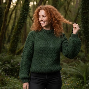 May include: A dark green textured knit sweater with a high neckline and long sleeves. The sweater is paired with black jeans. The person in the image is smiling and has red curly hair.