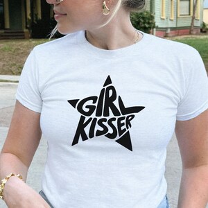 May include: White t-shirt with a black star graphic that reads "GIRL KISSER". The shirt has short sleeves and a crew neck. The model is wearing blue jeans and gold jewelry.