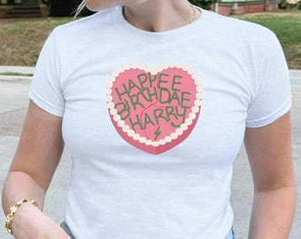 May include: White t-shirt with a heart-shaped graphic that reads "Happee Birthdae Harry" in pink and green. The shirt is short-sleeved and has a crew neck. The model is wearing blue jeans and gold jewelry.