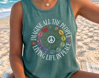 May include: Teal tank top with the text "IMAGINE ALL THE PEOPLE LIVING LIFE IN PEACE" encircling a peace sign. The text is in white, and the circle is decorated with colorful flowers. The person is sitting on a beach.