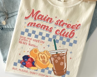 May include: Ivory-colored t-shirt with the text "Main Street Moms Club" in red and blue. The shirt features a graphic of a Mickey Mouse waffle, iced coffee, and berries. A light brown hat and blue jeans are also in the image.