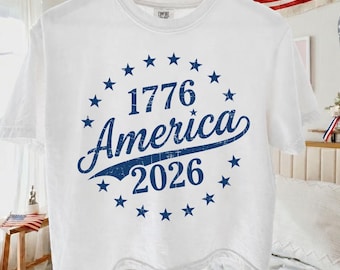 May include: White t-shirt with a blue graphic design. The design features the text "1776 America 2026" surrounded by blue stars. The shirt is hanging on a wooden hanger.