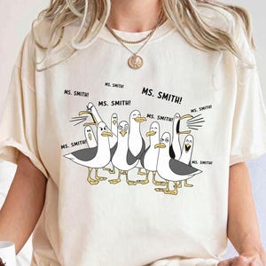 May include: Ivory-colored t-shirt featuring a cartoon of seagulls with the text "MS. SMITH!" above them. The shirt is made of a soft material and has a relaxed fit. The brand name "COMFORT COLORS" is printed on the top right.