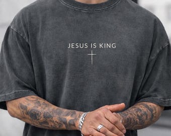 May include: Dark gray t-shirt with the text "JESUS IS KING" and a white cross. The shirt has a vintage, washed look. The short-sleeved shirt is worn by a person.