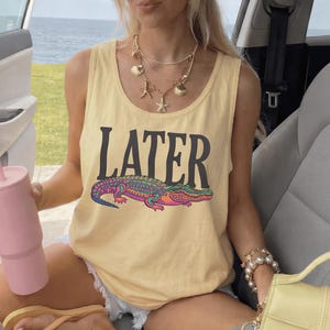 May include: A pale yellow sleeveless tank top featuring the word LATER in black capital letters above a colorful graphic of an alligator with purple, pink, and green patterns. The garment has a scoop neckline and is made of a solid cotton fabric.