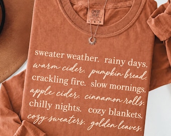 May include: A long-sleeve, rust-colored crewneck shirt featuring white text that reads: sweater weather, rainy days, warm cider, pumpkin bread, crackling fire, slow mornings, apple cider, cinnamon rolls, chilly nights, cozy blankets, cozy sweaters, golden leaves.