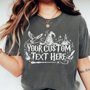 May include: A charcoal gray short-sleeve t-shirt featuring a white graphic print of a wizard hat, an owl, a candle, a potion bottle, and a wand. The text "YOUR CUSTOM TEXT HERE" is printed in the center in a stylized, jagged font.