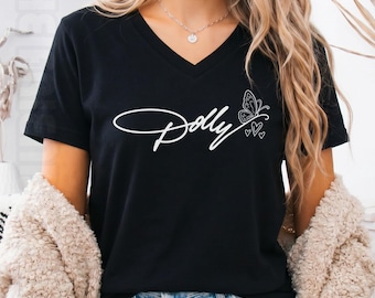 May include: A black V-neck t-shirt featuring the white cursive text Dolly with a butterfly and heart graphic on the chest. The shirt is worn with a beige textured cardigan and light blue denim shorts, showcasing a casual style.