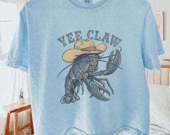 May include: Light blue t-shirt with a graphic of a lobster wearing a cowboy hat. The text "Yee Claw" is arched above the lobster. The shirt is hanging on a wooden hanger. The shirt is made of a soft material.