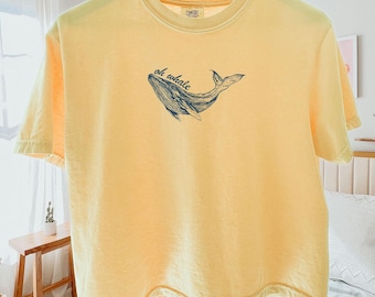 May include: Butter yellow t-shirt with a blue whale illustration and the text "oh whale". The short-sleeved shirt is made of a soft material and is hanging on a wooden hanger.