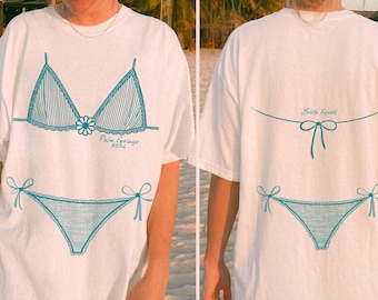 May include: White t-shirt with a teal bikini graphic on the front and back. The front design includes a bikini top and bottom with the text "Palm Springs 2026". The back of the shirt has a bikini graphic and the text "Bride Squad".