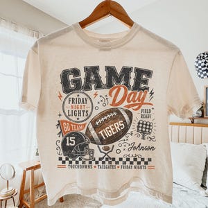May include: An ivory short-sleeve t-shirt featuring a graphic print with the text GAME DAY, FRIDAY NIGHT LIGHTS, GO TEAM, TIGERS, and JOHNSON. The design includes a football, a helmet with the number 15, a scoreboard, and a checkered border.