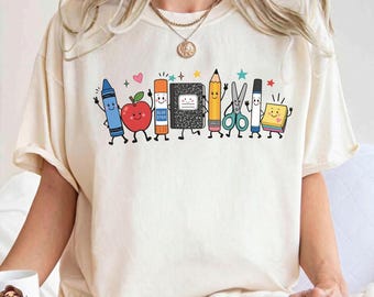 May include: An ivory short-sleeve t-shirt featuring a graphic print of cartoon school supplies including a blue crayon, red apple, glue stick, black notebook, yellow pencil, scissors, marker, and sticky notes with small colorful stars and hearts.