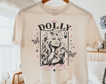 May include: An ivory short-sleeve t-shirt featuring a black and pink graphic print of Dolly Parton in a playing card frame. The design includes the text DOLLY, the letter Q, and butterfly accents. The shirt is displayed on a wooden hanger.