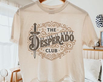 May include: An ivory short-sleeve t-shirt featuring a brown graphic print of a sword and ornate scrollwork. The text reads THE DESPERADO CLUB and SO FUN IT HURTS. The shirt is displayed on a wooden hanger against a light-colored bedroom background.