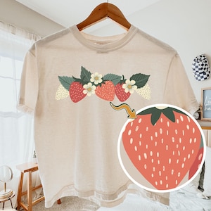 May include: An ivory short-sleeve t-shirt featuring a graphic of red strawberries and white flowers across the chest. A circular inset shows a magnified view of a single strawberry with a yellow arrow pointing to it. The text COMFORT COLORS is visible.