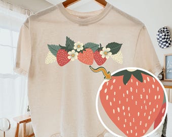 May include: An ivory short-sleeve t-shirt featuring a graphic of red strawberries and white flowers across the chest. A circular inset shows a magnified view of a single strawberry with a yellow arrow pointing to it. The text COMFORT COLORS is visible.