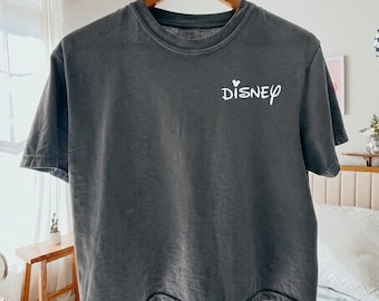 May include: A dark gray t-shirt with the word "Disney" in white script. The shirt is hanging on a wooden hanger. The shirt is a Comfort Colors brand.