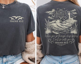 May include: A charcoal gray t-shirt featuring a small graphic on the front with three birds and the text Isaiah 43:2. The back displays a large illustration of a lighthouse, crashing waves, birds, and the text When you go through deep waters I will be with you Isaiah 43:2.