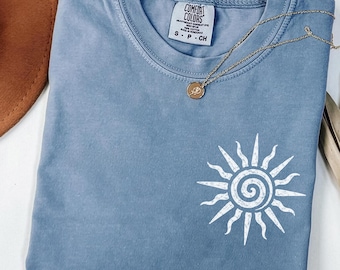 May include: A blue jean colored t-shirt with a white sun graphic on the front. The shirt has a crew neck and short sleeves. A gold necklace with a pendant is draped across the shirt. The tag reads "Comfort Colors".