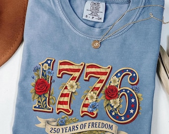 May include: A light blue Comfort Colors t-shirt featuring the number "1776" in red, white, and blue, with floral accents and the text "250 YEARS OF FREEDOM" on a banner. A gold necklace with a pendant is draped across the shirt.