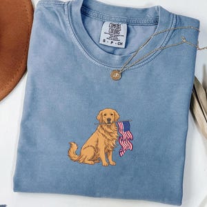 May include: A blue t-shirt with a graphic of a golden retriever holding an American flag. The shirt has a crew neck and short sleeves. A gold necklace with a pendant is draped on the shirt.