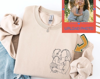 May include: Beige sweatshirt with a black line drawing of a mother and child. A small photo with the text "UPLOAD PHOTO" is in the upper right corner. The sweatshirt is folded on top of a pair of blue jeans.