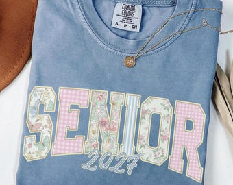 May include: A blue pigment-dyed t-shirt featuring the word SENIOR in large block letters with alternating floral and pink gingham patterns, with the year 2027 printed below in a light blue script font and a gold-toned pendant necklace resting on top.