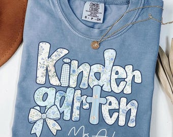 May include: A blue jean colored t-shirt with a crew neckline and a Comfort Colors label. The front features the text Kindergarten in a floral and gingham pattern, a floral bow, and the name Mrs. Anderson in white script lettering.