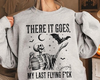 May include: A light gray crewneck sweatshirt featuring a black graphic print of a skeleton holding a birdcage and reaching toward a flying bat. The text reads, "THERE IT GOES, MY LAST FLYING F*CK," surrounded by stars and a crescent moon design.