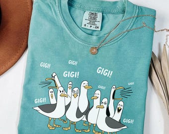 May include: Seafoam green t-shirt with a crew neck and short sleeves. The shirt features a cartoon illustration of seagulls with the word "GIGI!" repeated around them. A gold necklace is draped on the shirt.